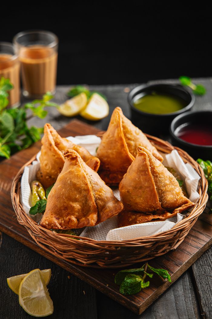 services-04 Fried golden samosas on white tissues in wicker basket on board surrounded by lemon and salad dressing in deep dishes and beverages in glass